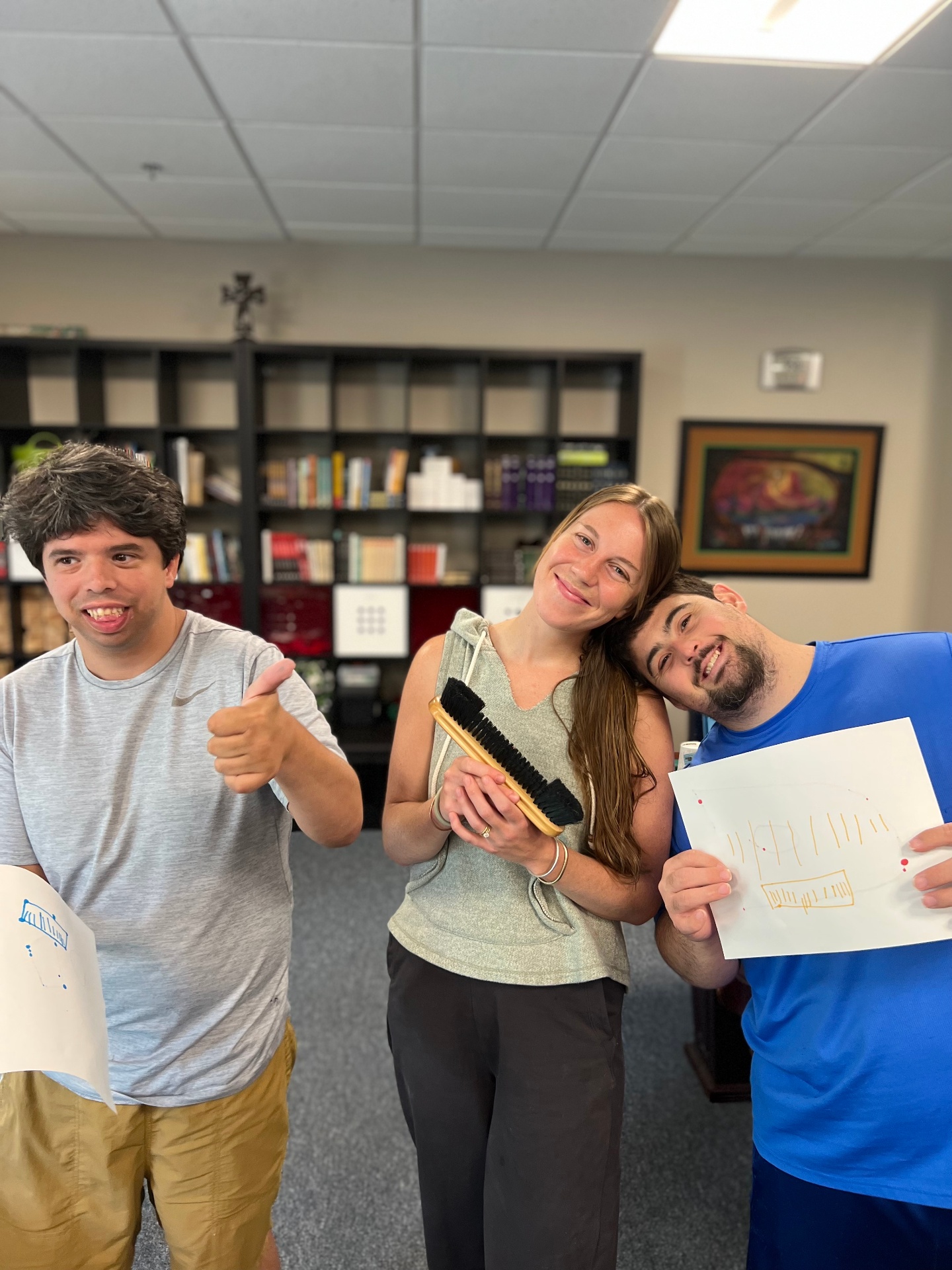 An occupational therapist smiling alongside two young adult clients proudly holding up their artwork during a vocational skills session